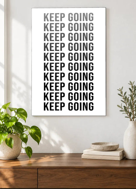 Keep Going in Full Focus acrylic wall art displayed above a wooden console with plants and books against a white wall.