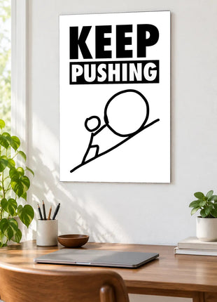 Keep Pushing Uphill acrylic wall art displayed above a wooden desk with a laptop, plants and books against a white wall.