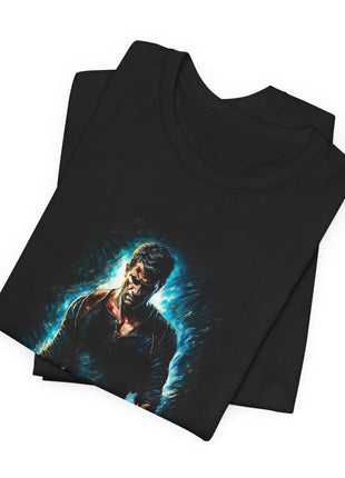 Folded black T-shirt close-up showing Nathan Drake’s face, blue shirt and shoulder straps framed by bright cyan and white brushwork.