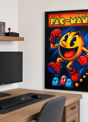 Pac-Man wearing red gloves and boots above three small ghosts, with arcade lettering across the top on acrylic wall art beside a computer monitor and oak desk in a game room with white walls.