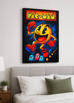 Pac-Man wearing red gloves and boots above three small ghosts, with arcade lettering across the top on canvas wall art above a gray upholstered headboard in a bedroom with white bedding.