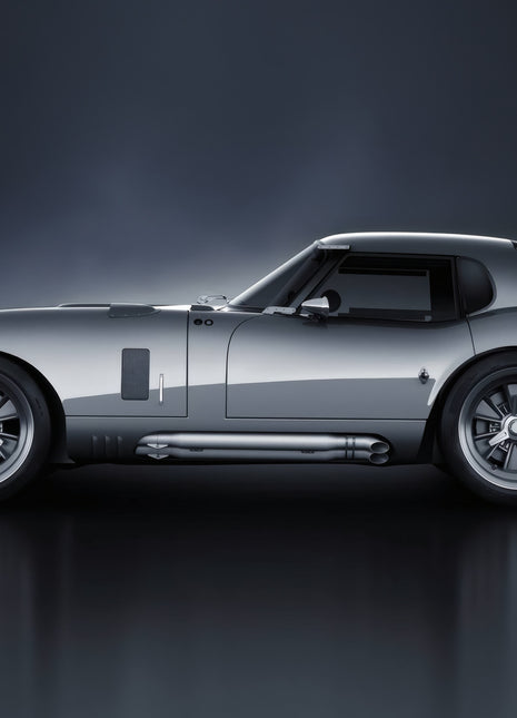 Silver long-nose racing coupe facing left in side profile, with a fastback roof, rear lip spoiler and side exhaust over a gray reflection.