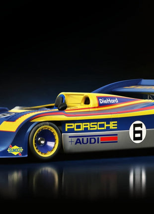Blue and yellow number 6 Porsche prototype racer with an open cockpit, tall rear wing, yellow wheel rims and Porsche Audi lettering on a dark floor.