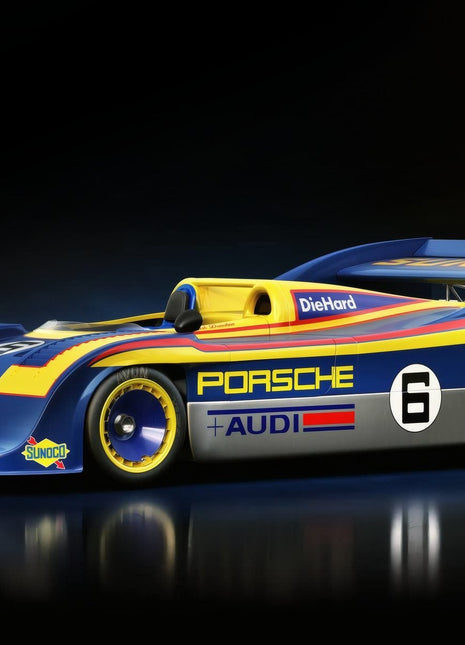 Blue and yellow number 6 Porsche prototype racer with an open cockpit, tall rear wing, yellow wheel rims and Porsche Audi lettering on a dark floor.