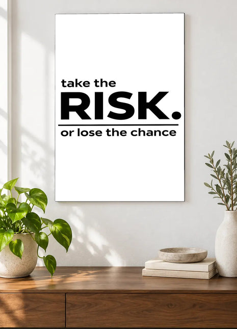 Take the Risk acrylic wall art displayed above a wooden console with plants and books against a white wall.