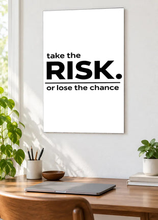 Take the Risk acrylic wall art displayed above a wooden desk with a laptop, plants and books against a white wall.